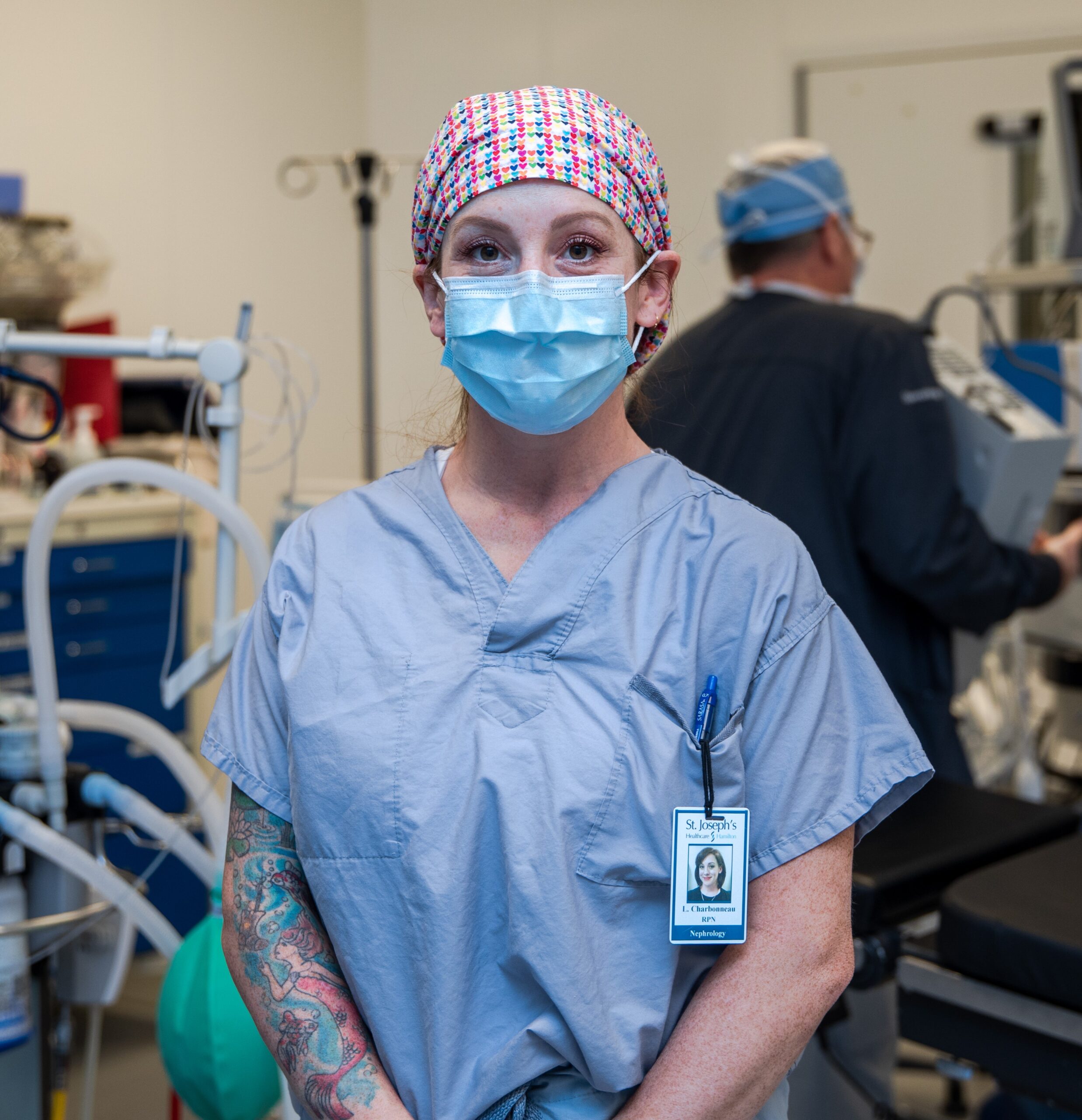 Registered Practical Nurse Charbonneau stands in the operating room at St. Joseph’s Healthcare Hamilton surrounded by operating room equipment. Charbonneau is wearing a surgical cap, mask and scrubs, and is looking directly at the camera with hands clasped gently in front of her.