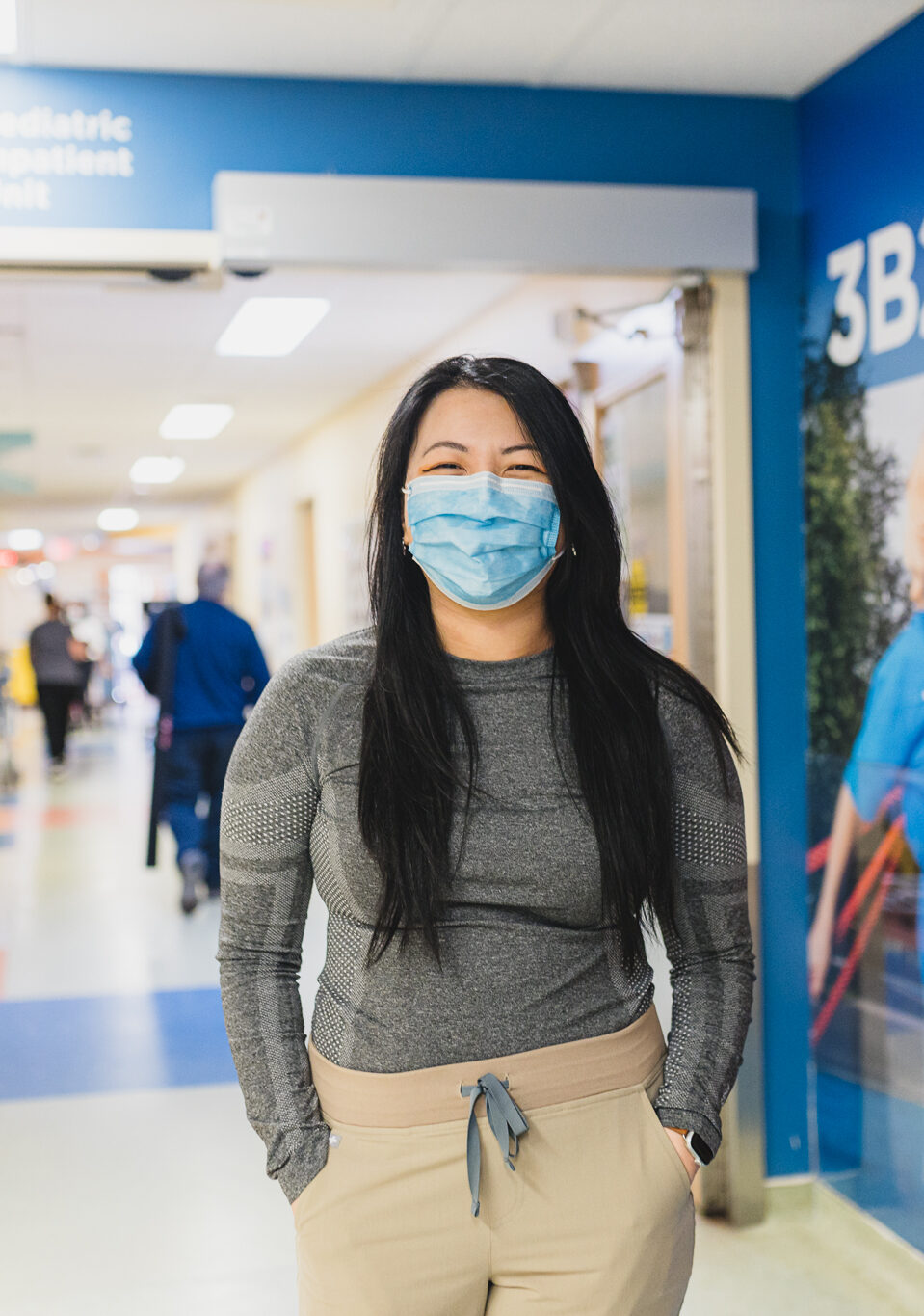 Registered Nurse Tran Thai works in the pediatric oncology department at Hamilton Health Sciences McMaster Children's Hospital. She is standing in the hospital hallway, in front of colourful wall murals. She has long dark hair and is wearing a medical mask.